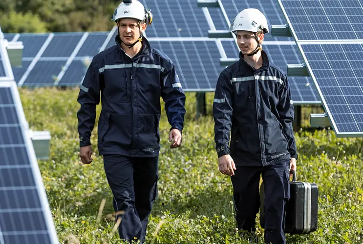 Two men in dark blue protective clothing and white helmets walk with a toolbox through a meadow field on which PV systems have been installed.