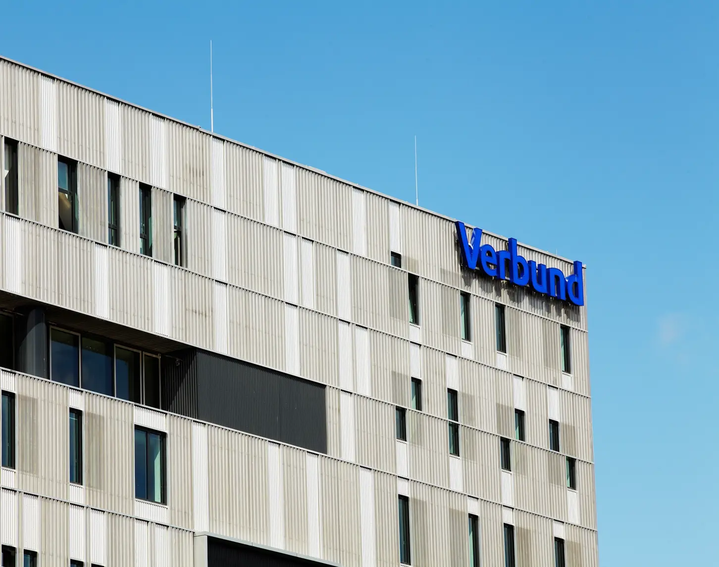 We look at the façade of VERBUND at Westbahnhof. It is grey with windows. At the top right is the VERSBUND lettering in VERBUND blue.